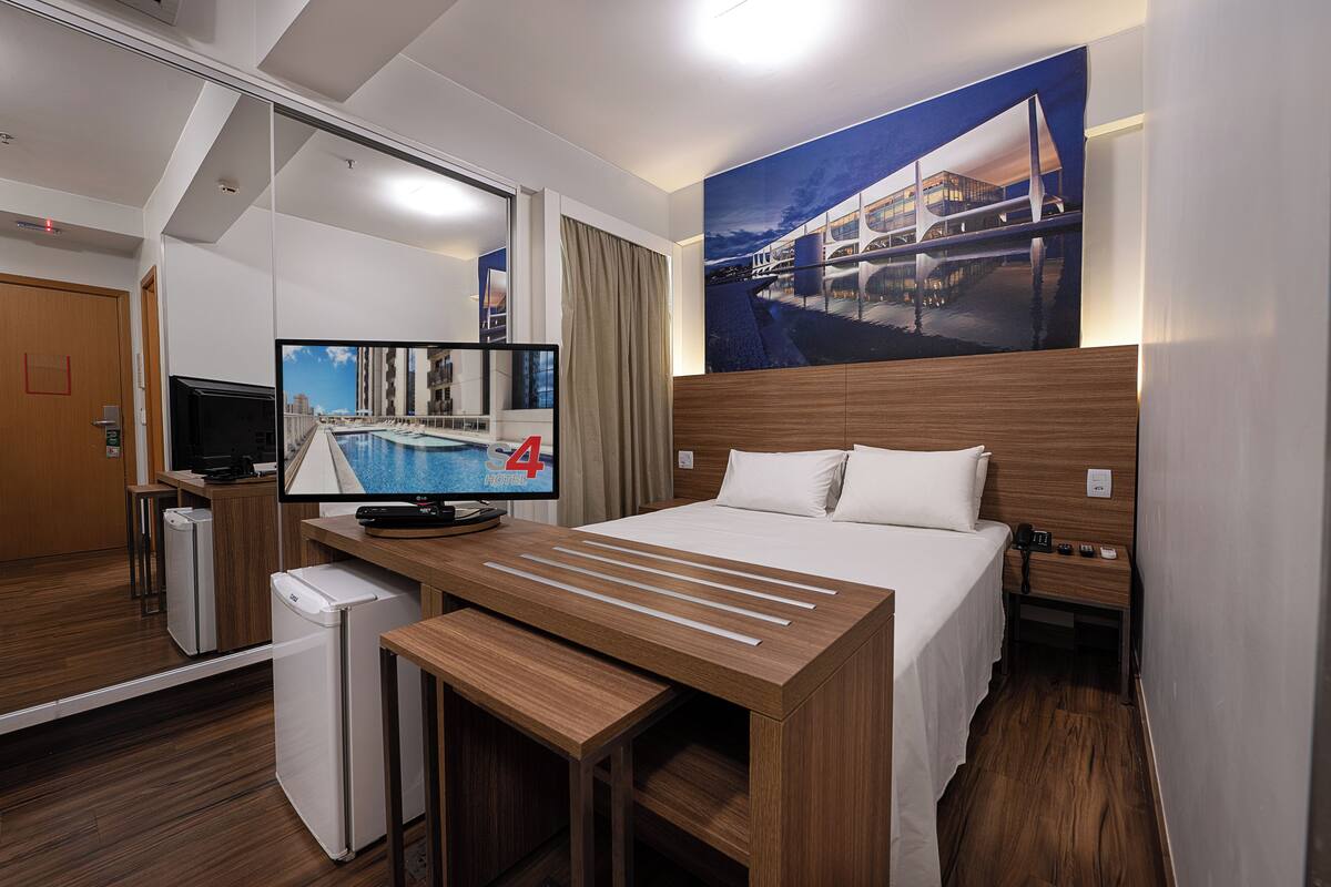 Quarto de hotel com uma cama, escrivaninha com computador, televisão e mini-geladeira.