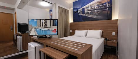 A hotel room with a bed, a desk with a computer, a television, and a small refrigerator.