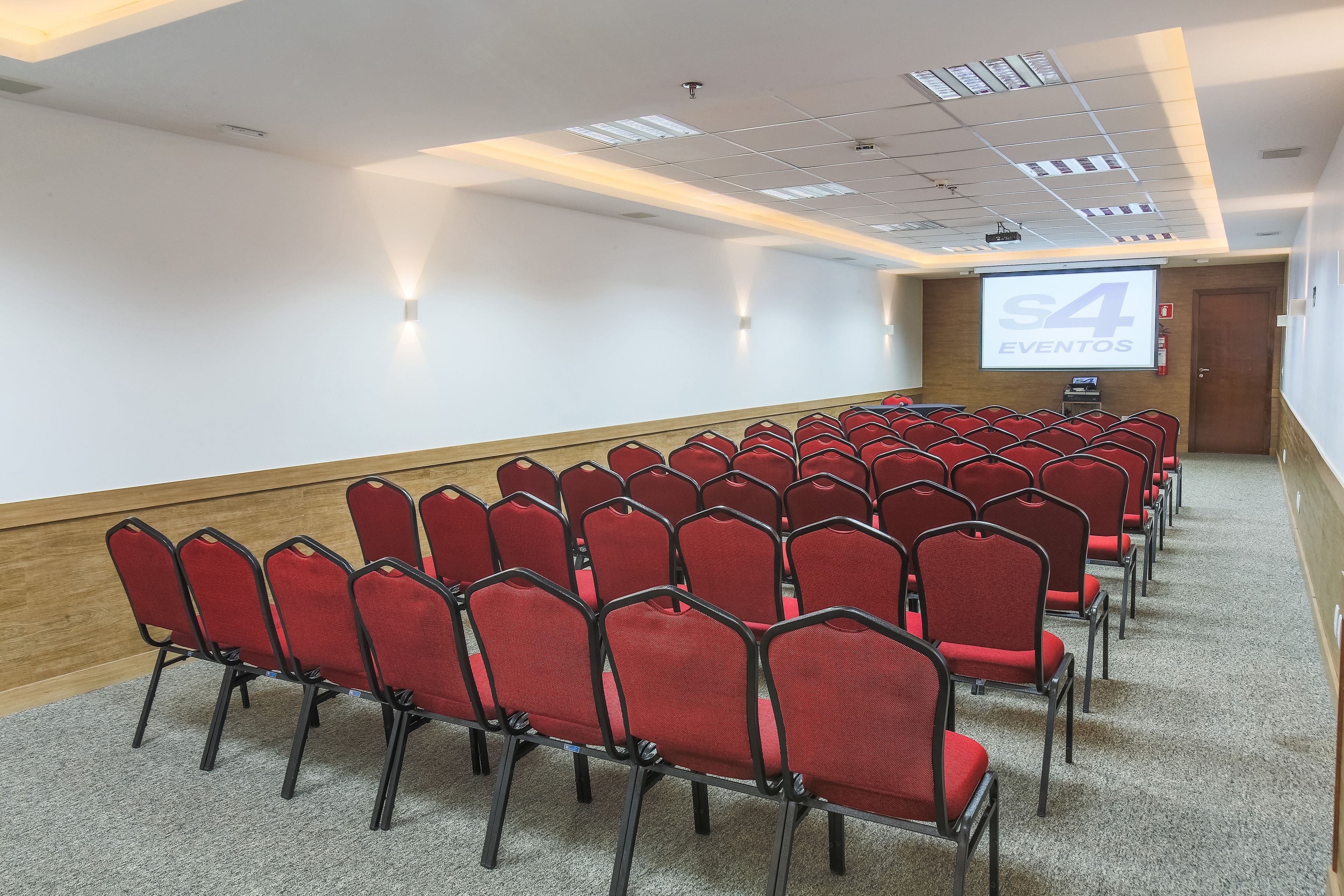 A conference room with rows of red chairs, a projector screen displaying "S4 EVENTOS", and a wooden accent wall.