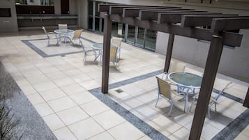 Uma área externa ampla com várias mesas e cadeiras redondas, uma estrutura de pérgola e piso revestido de azulejos.