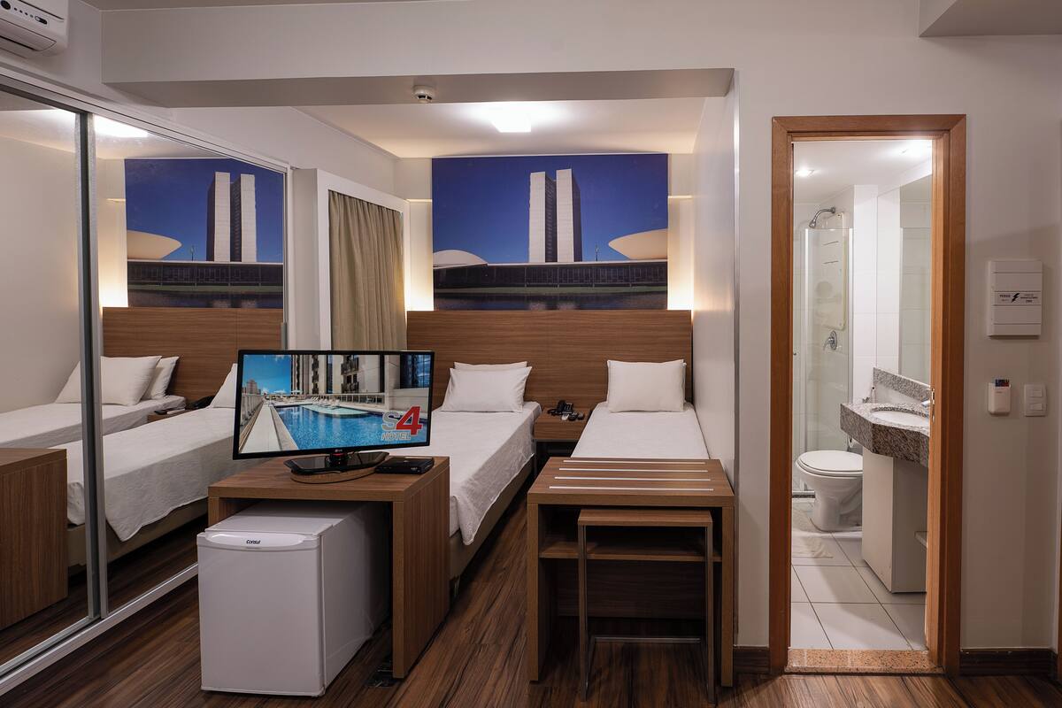 Quarto de hotel com duas camas, uma mesa de escritório com computador e geladeira, uma pintura grande de uma ponte e banheiro com espelho e vaso sanitário.