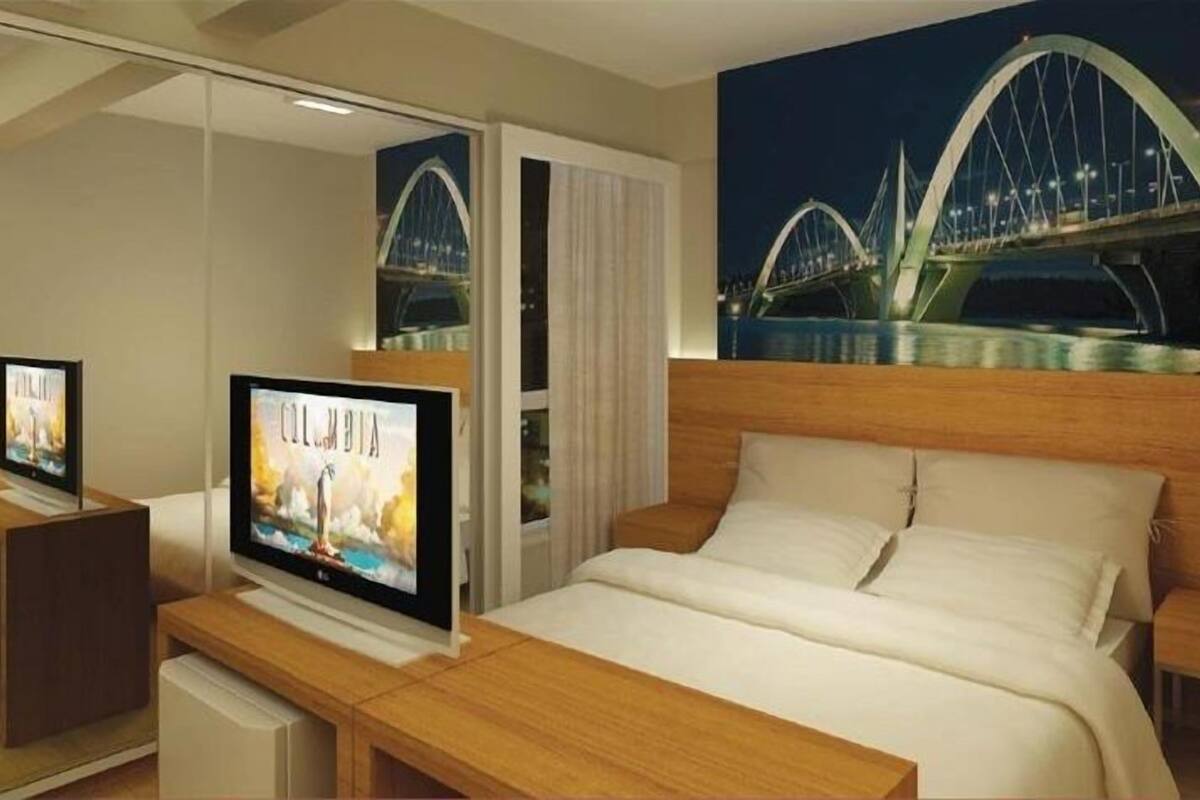 Quarto de hotel com cama, TV sobre apoio de madeira e um espelho grande refletindo uma ponte à noite.