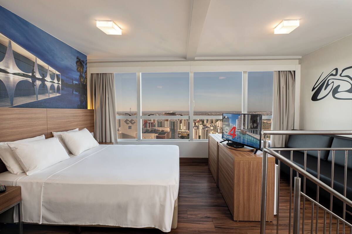 Quarto de hotel com uma cama grande, uma escrivaninha com computador e vista para a cidade através de amplas janelas.