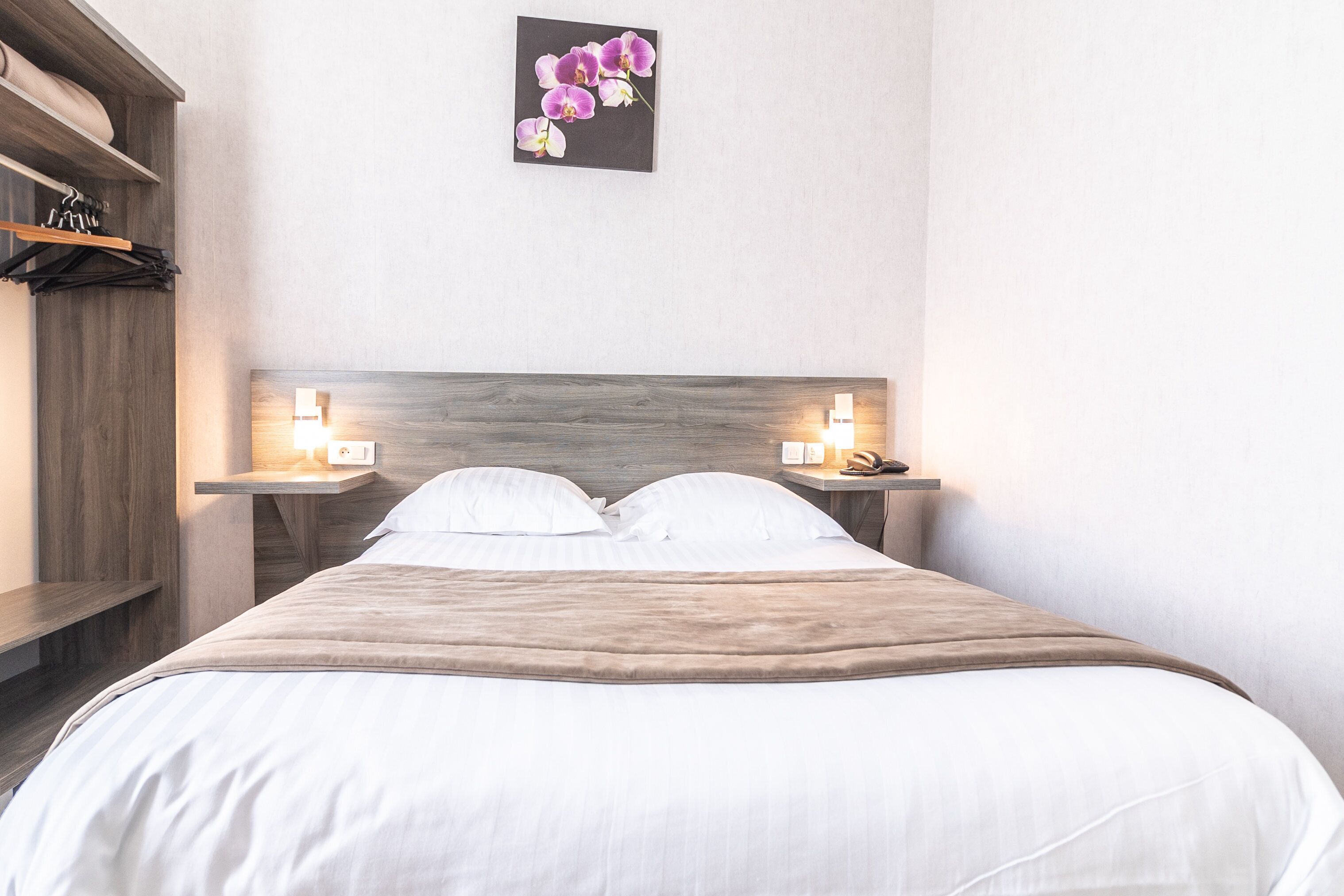 A neatly made bed with white linens and a brown throw blanket, flanked by two bedside tables with lamps. A wooden headboard and a wall-mounted picture of purple orchids are also visible.