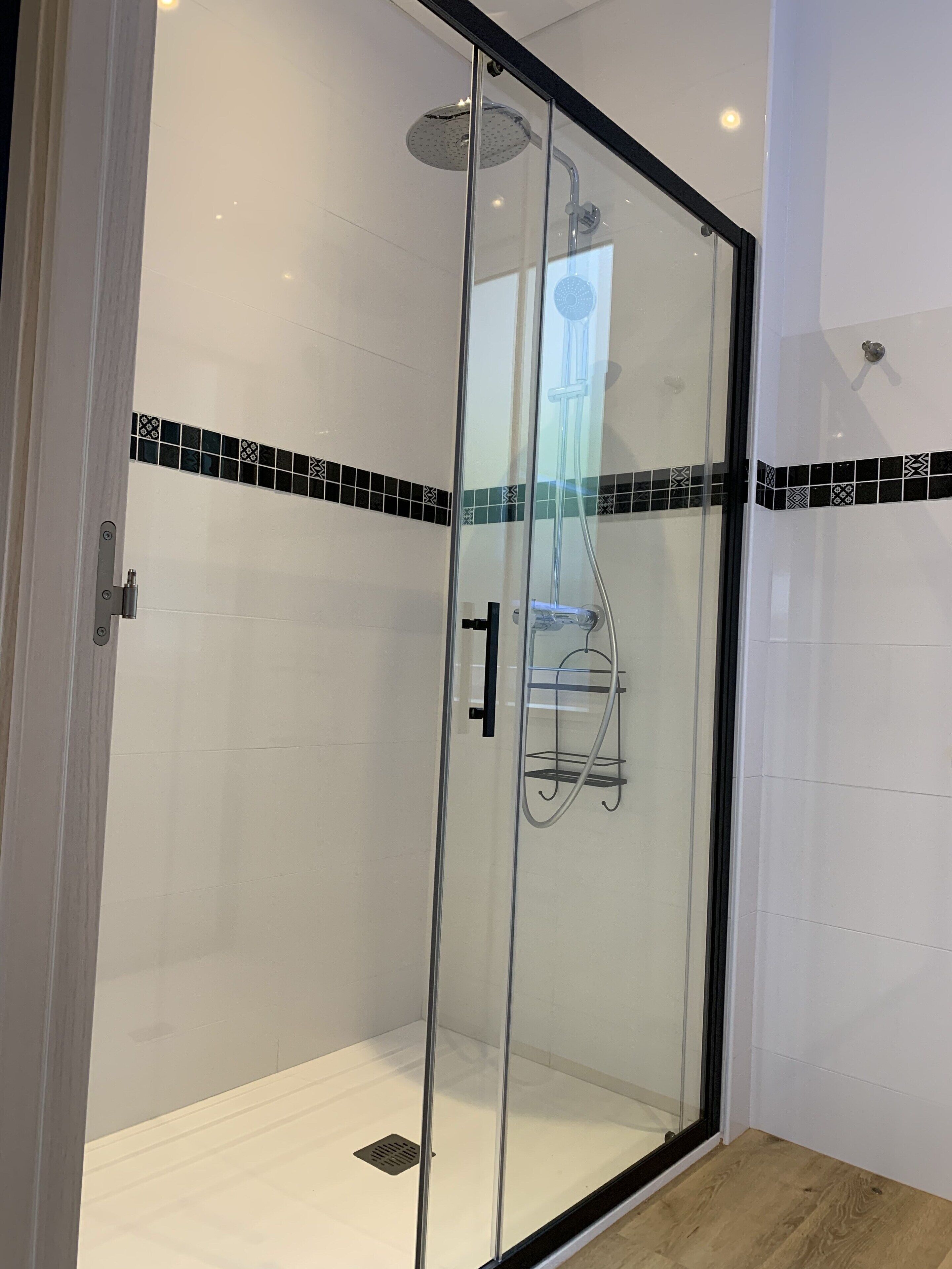 Ducha con mampara de vidrio, borde de azulejos blanco y negro, cabezal de ducha y pasamanos.