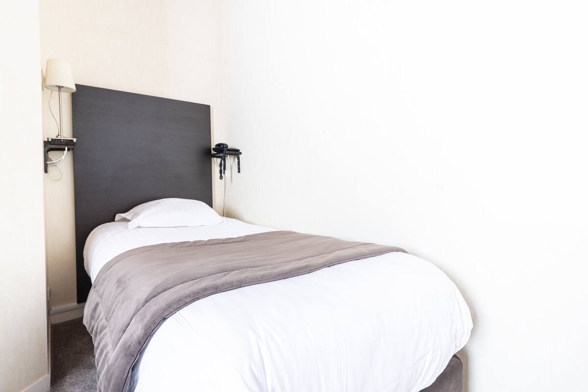 Een net opgemaakt bed met een donker hoofdbord, witte kussens en een grijze deken.