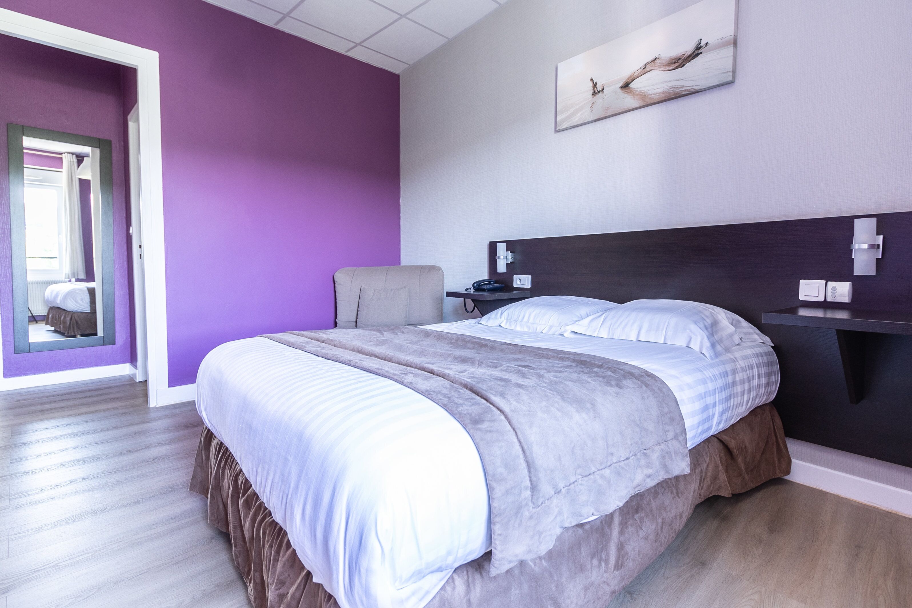 A modern hotel room with a large bed, a chair, a bedside table, and a painting on the wall.