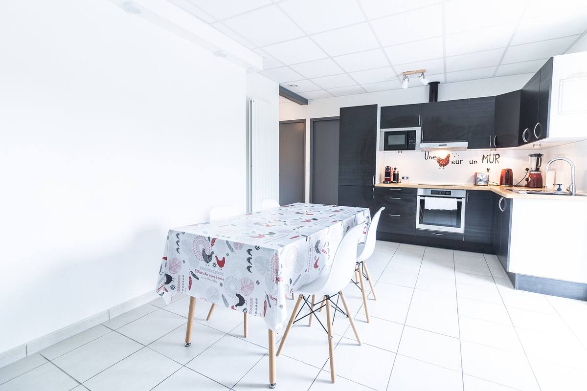 Een moderne keuken met donkere kastjes, een magnetron en een koffieapparaat. Er is een eettafel met stoelen en een vloer van witte tegels.