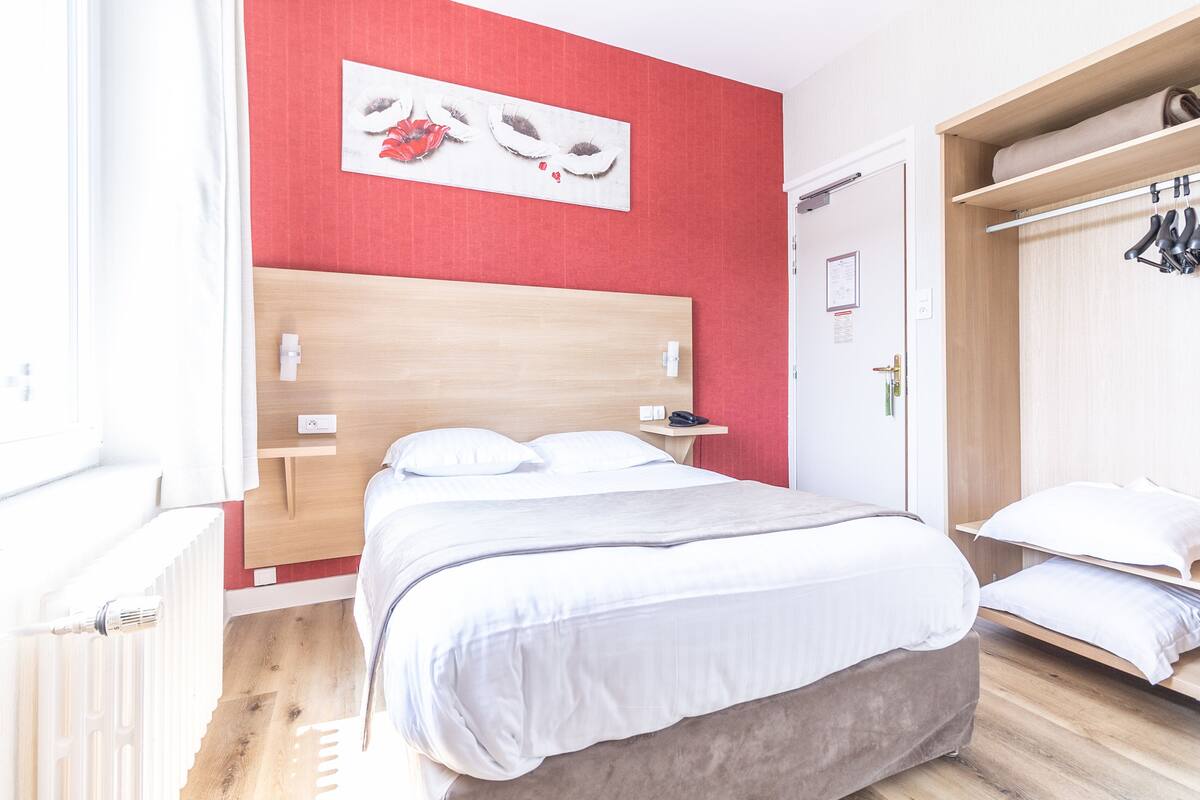Een slaapkamer met een houten hoofdeinde, een bed met witte beddengoed, een rode accentmuur en een houten garderobe met kledingroede.