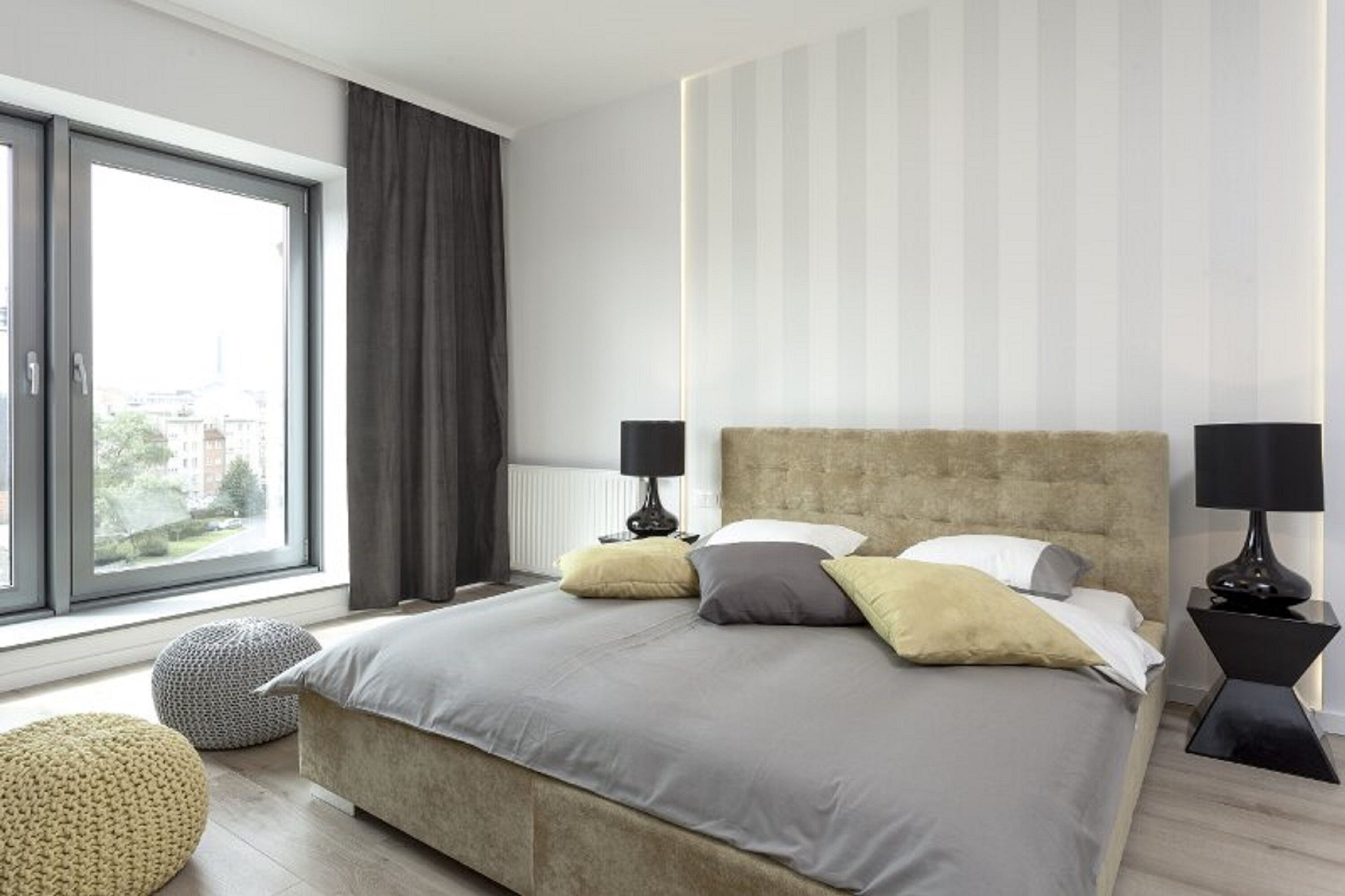 A modern bedroom with a large bed, two bedside tables, a window with curtains, and a view of buildings outside.