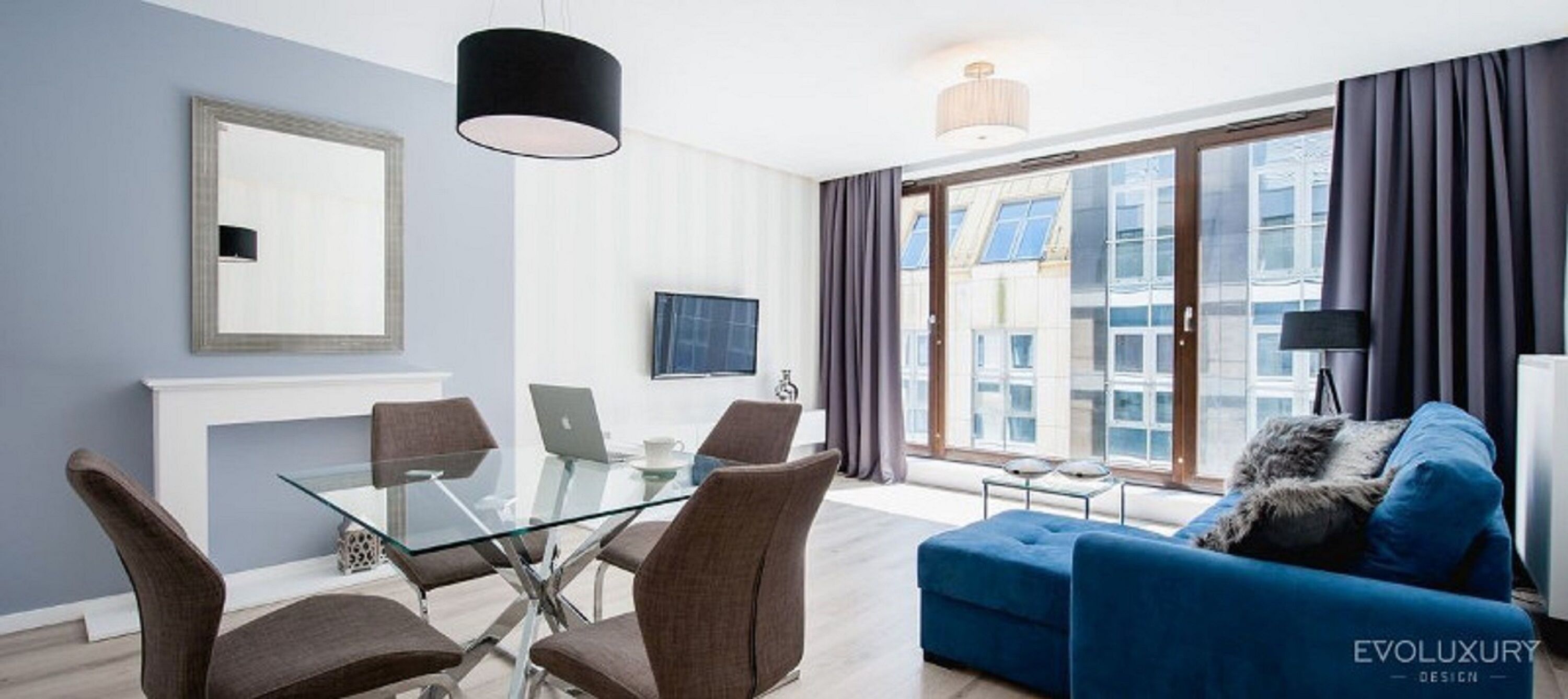 A modern living room with a glass dining table, brown chairs, a blue sofa, and a TV on the wall.