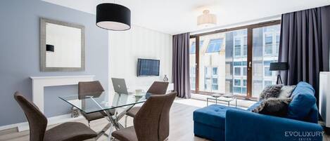 A modern living room with a glass dining table, brown chairs, a blue sofa, and a TV on the wall.