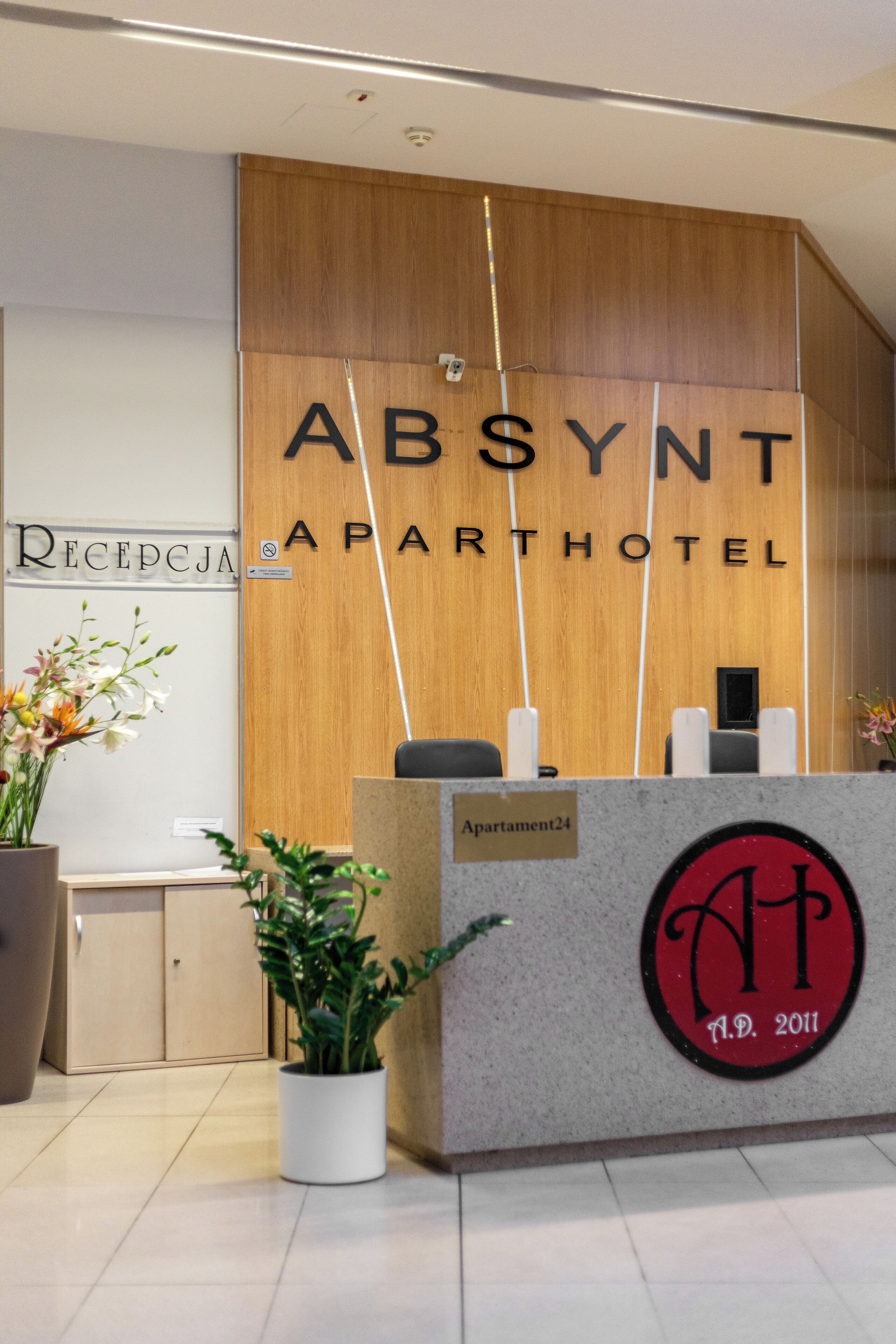 A reception desk with a wooden panel and a sign that reads "ABSYNT APARTHOTEL." There is a potted plant to the left of the desk and a sign indicating "Apartment24."