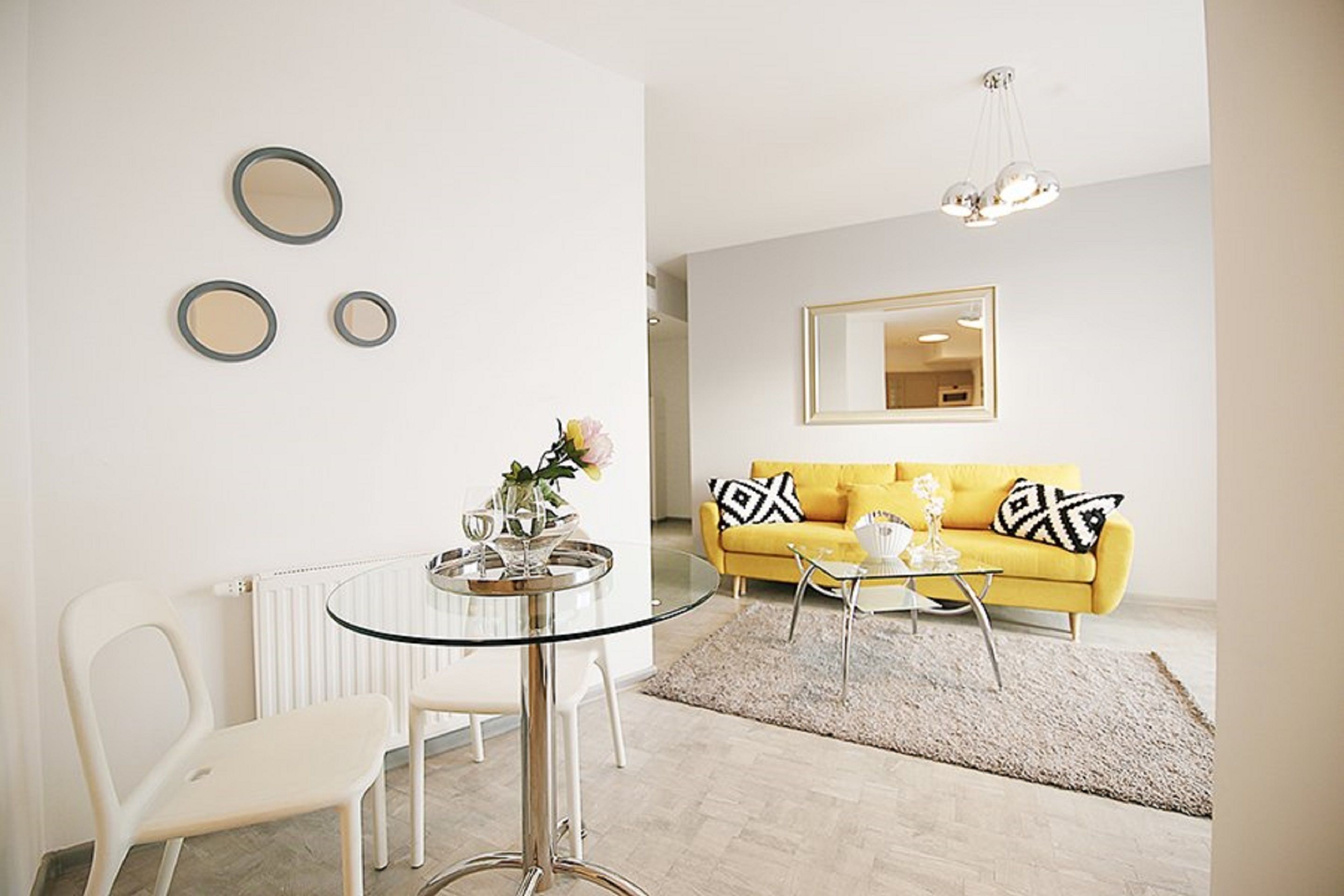 A modern living room with a glass table, white chairs, a yellow sofa, and a large mirror on the wall.