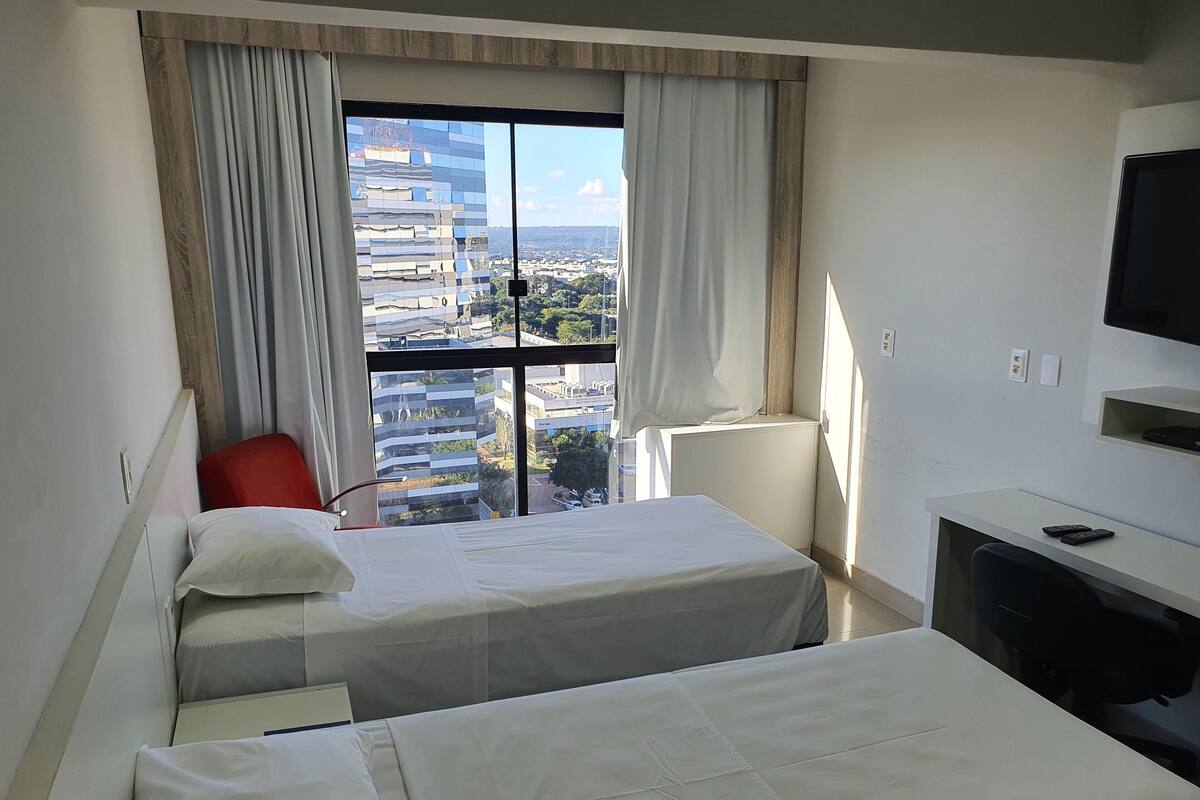 Habitación de hotel con dos camas, un escritorio, una silla, un televisor y ventana con vista a la ciudad.