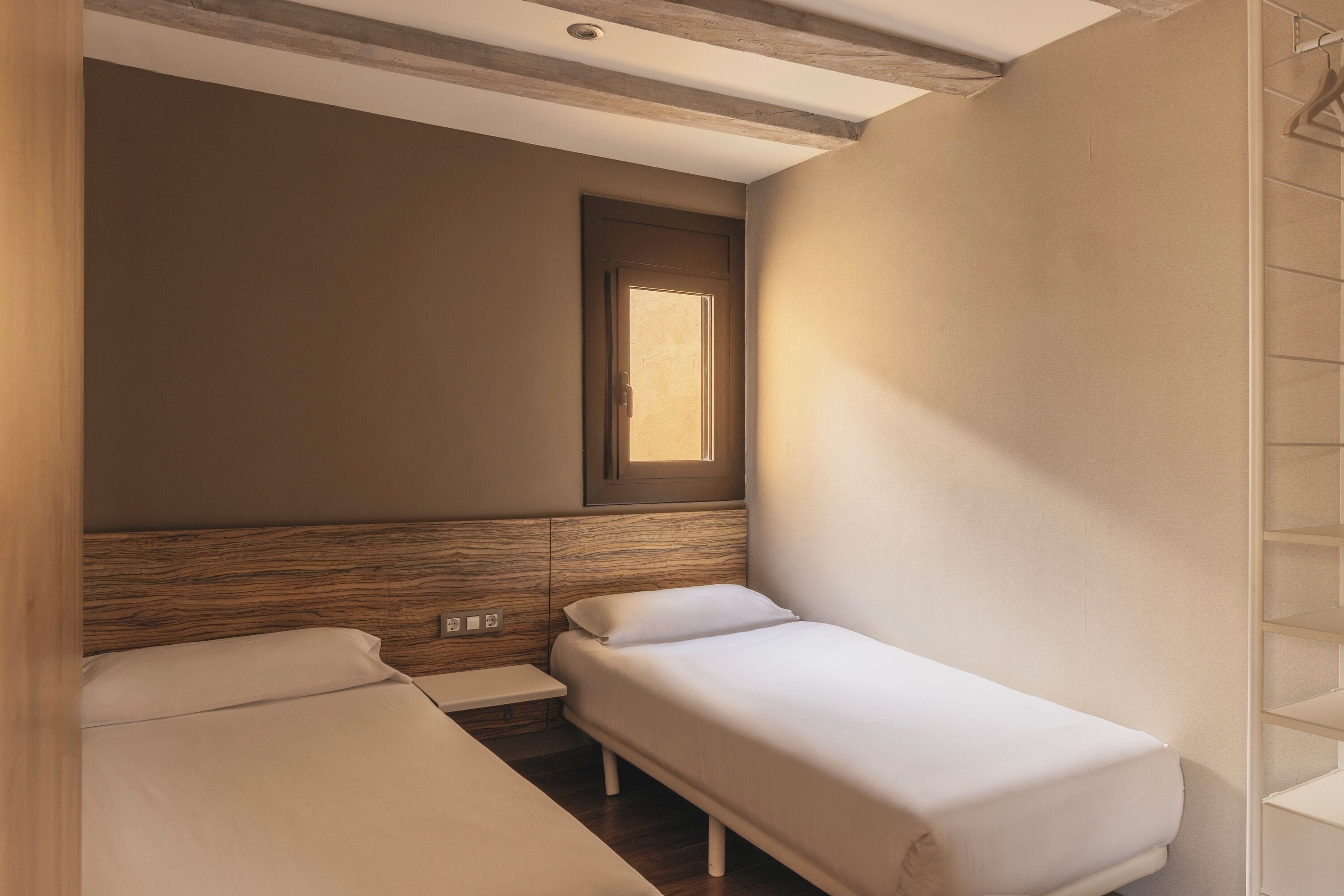 A small, single-room hotel bedroom with a wooden headboard, a window, and a built-in shelf.