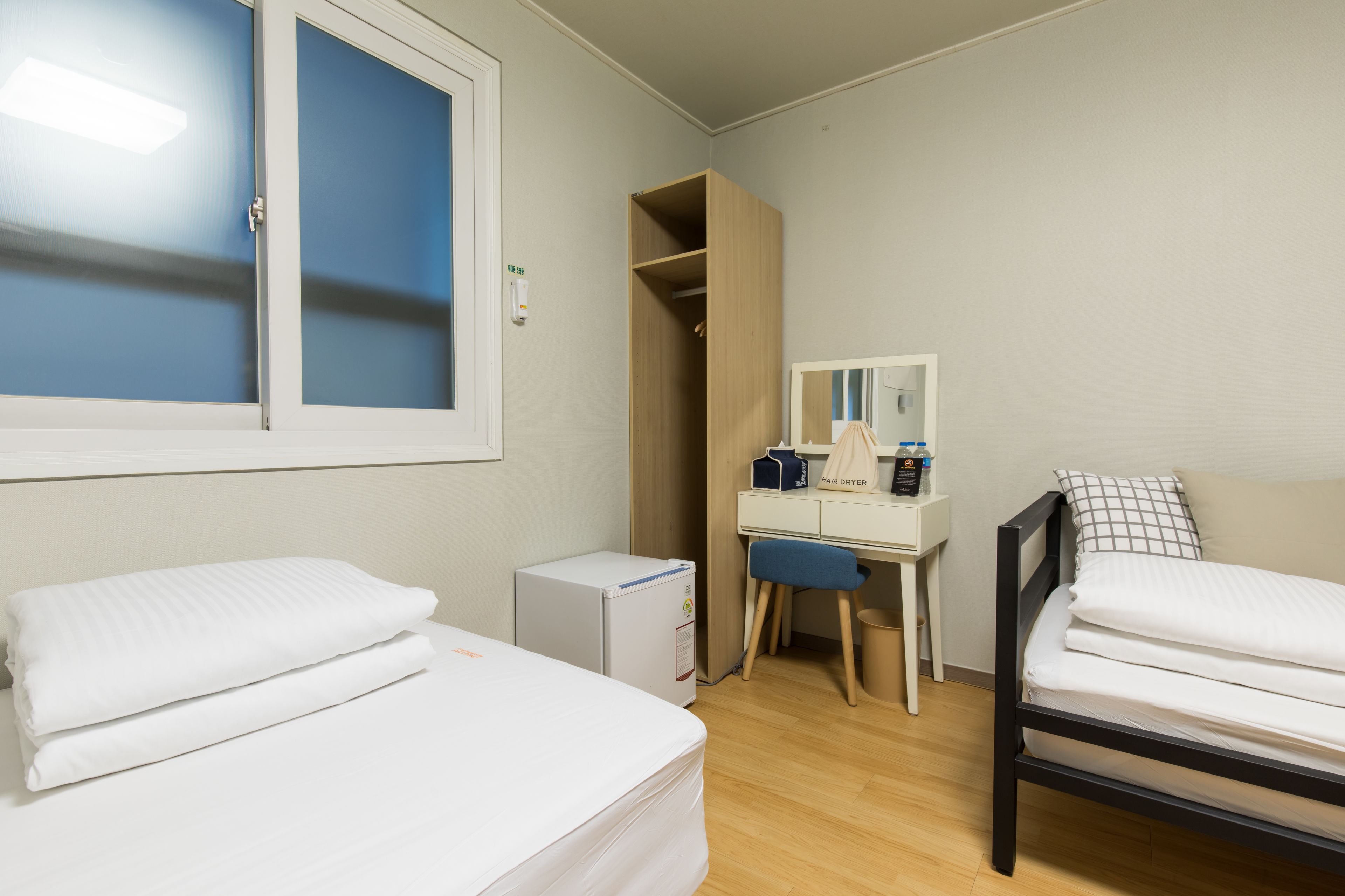A bedroom with two beds, a desk, a wardrobe, and a small refrigerator.