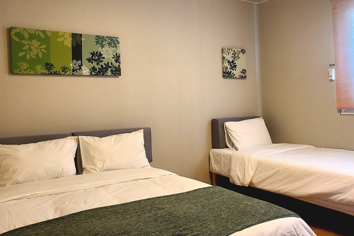 A room with two beds, each with white bedding and a green throw blanket. There are two floral wall art pieces above the beds.