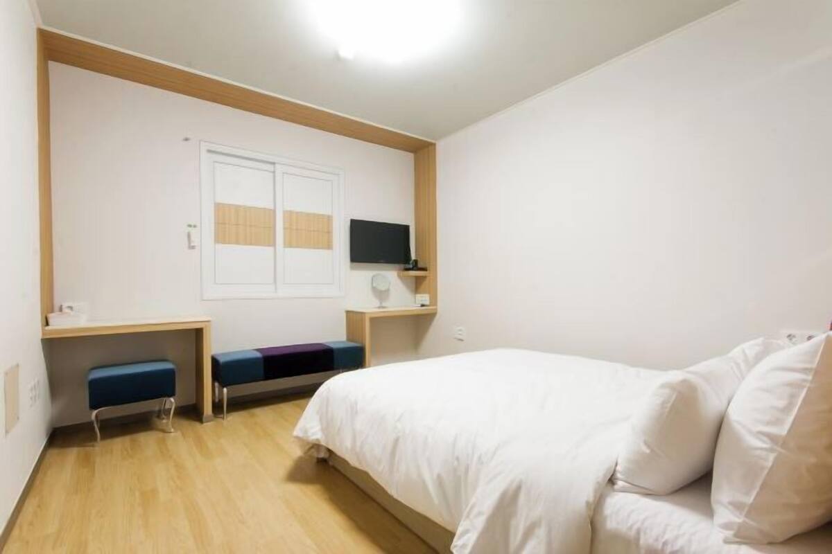 A bedroom with a bed, a desk, a chair, a television, and a window.