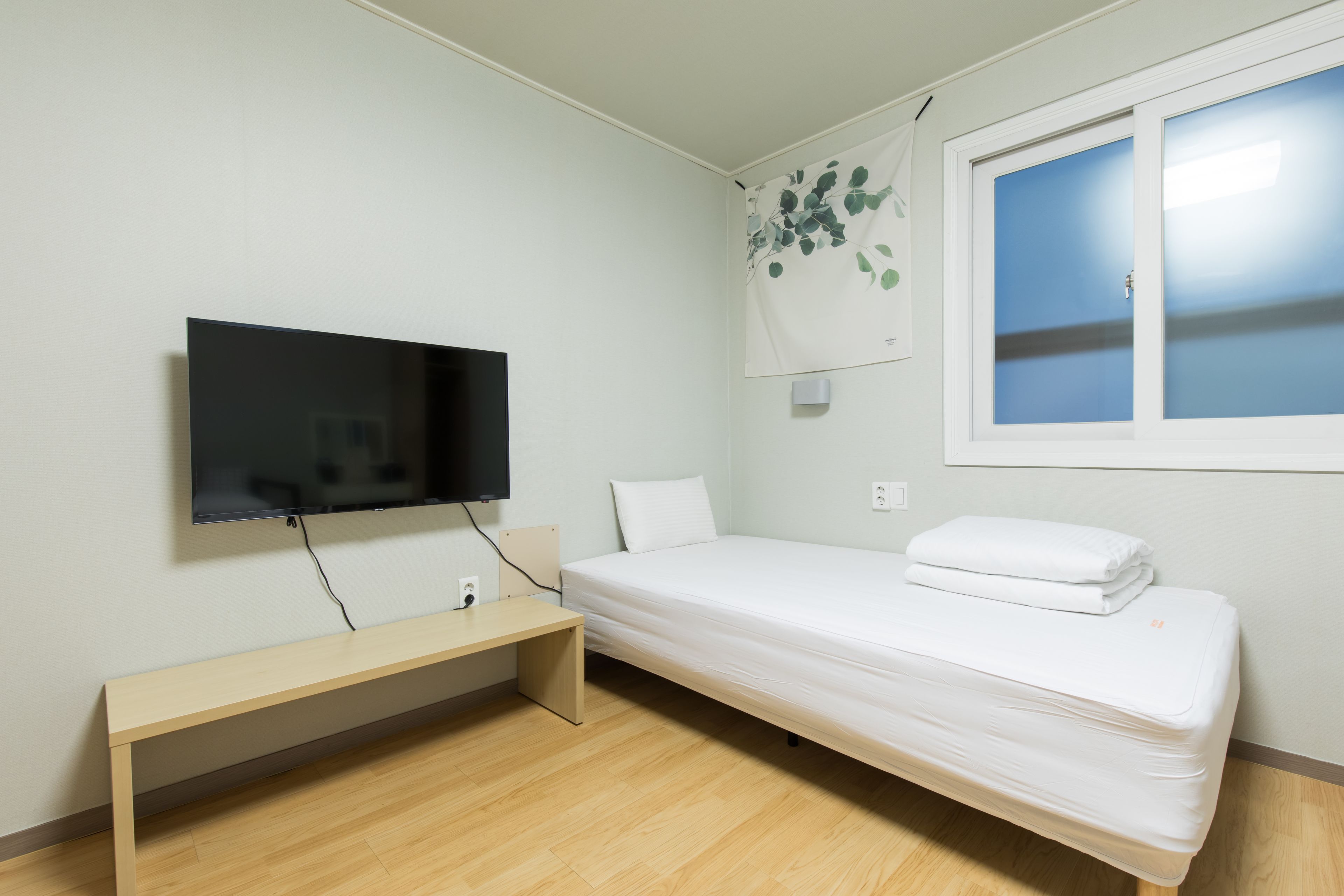 A small, modern room with a bed, a TV mounted on the wall, a bench, and a window with a view.
