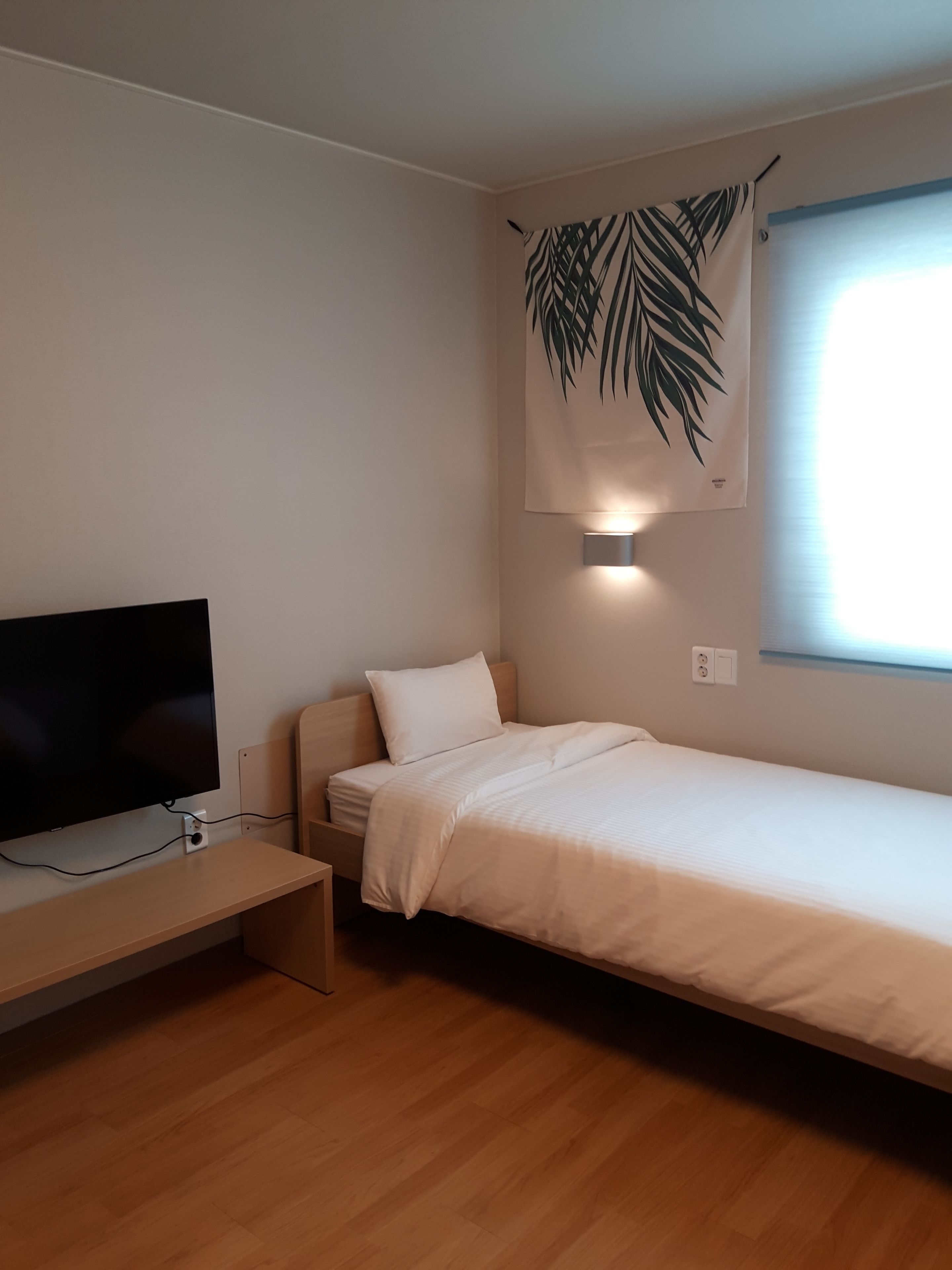 A small, well-lit room with a bed, a TV, and a wall-mounted lamp.