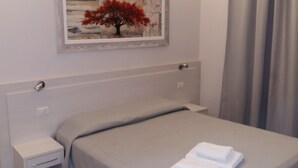 Cama bem arrumada com um espalha cama cinza e uma toalha branca dobrada por cima. Há uma mesa de cabeceira com uma gaveta e uma luminária pequena. Um quadro emoldurado com uma árvore e folhas vermelhas está pendurado na parede acima da cama.