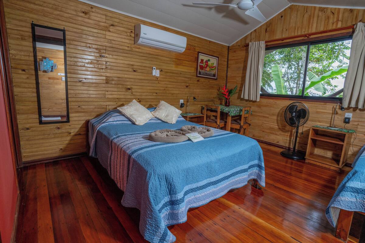 A wooden-paneled room with a bed, a desk, a chair, a fan, and a window with a view of trees.