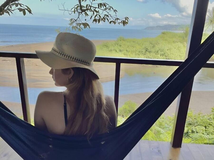 A person relaxing in a hammock on a wooden deck with a view of the beach and ocean.