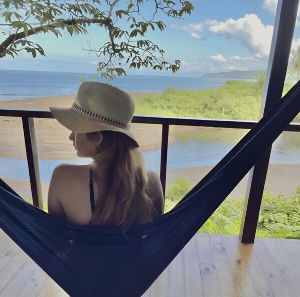 A person relaxing in a hammock on a wooden deck with a view of the beach and ocean. - Pirate Cove (Drake Bay)