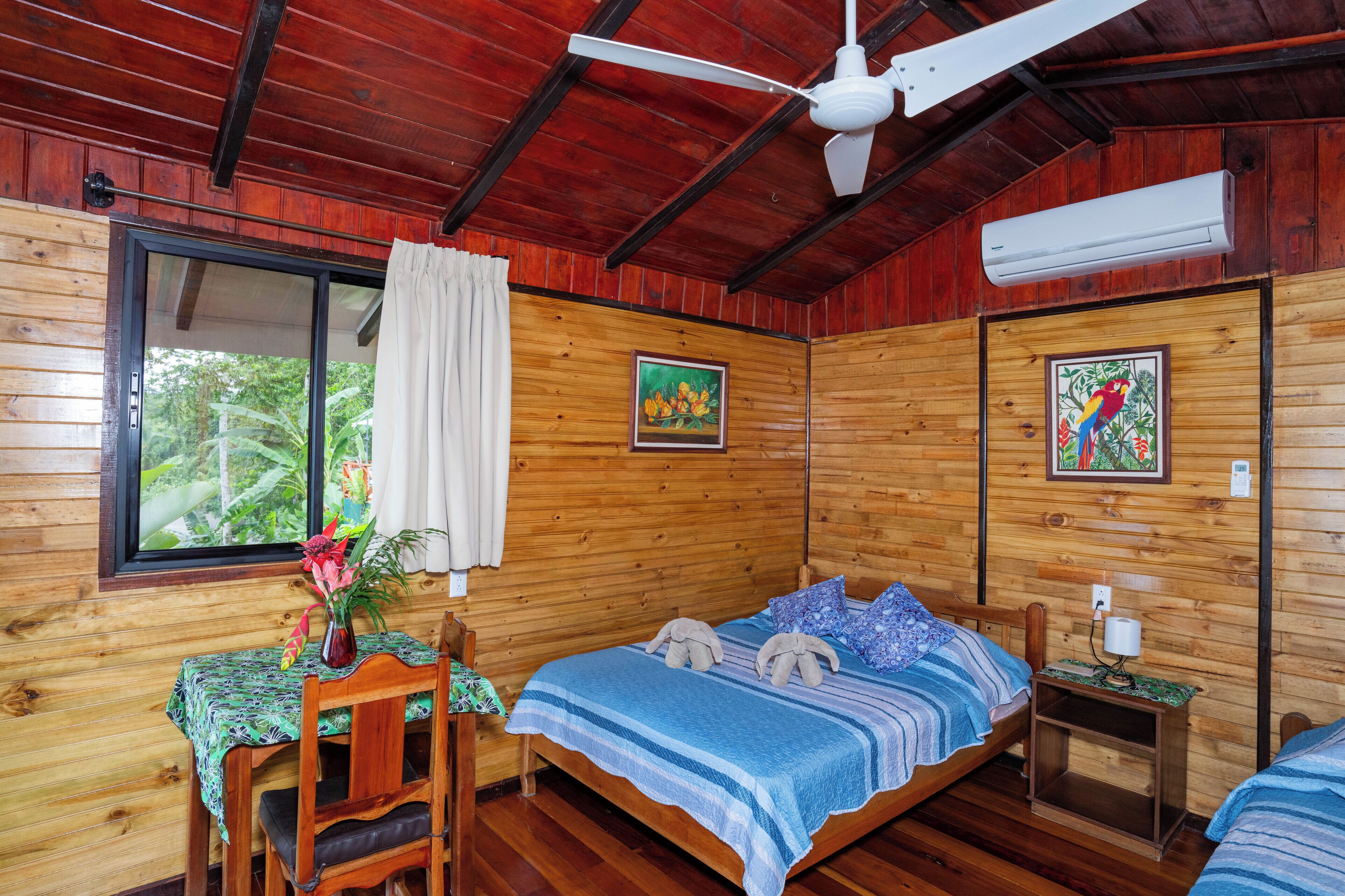 A wooden cabin room with a bed, a desk with a chair, a window with curtains, and two framed pictures on the walls.