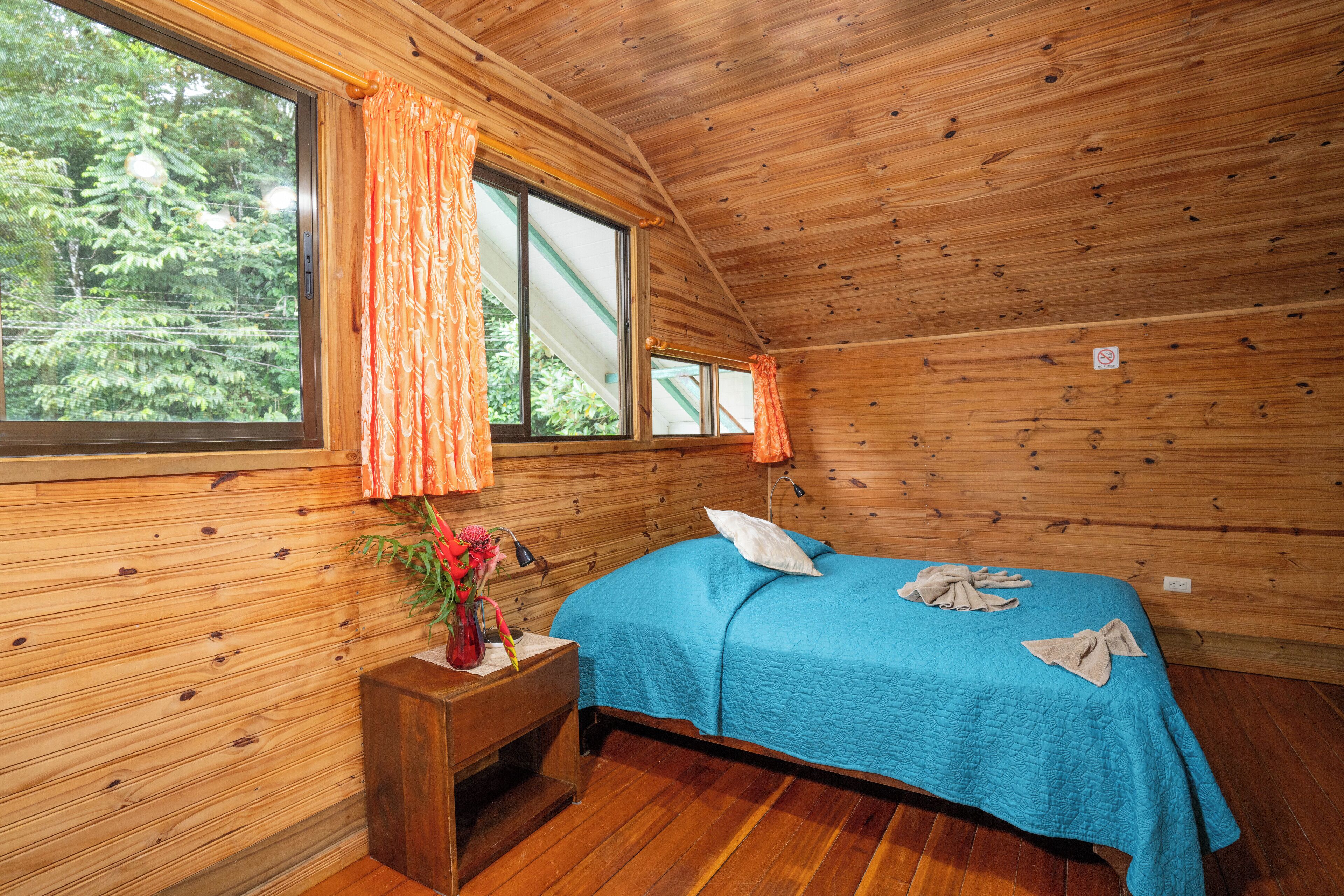 A wooden cabin room with a bed, a nightstand, a window with curtains, and a view of trees.