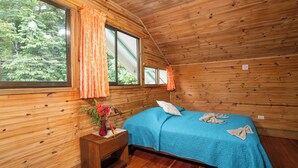 A wooden cabin room with a bed, a nightstand, a window with curtains, and a view of trees. - Pirate Cove (Drake Bay)
