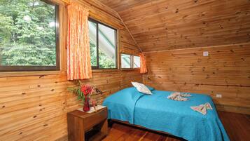 Habitación de cabaña de madera con cama, mesita de noche, ventana con cortinas y vista a árboles.