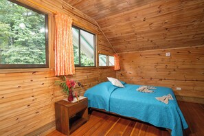 Quarto de cabana de madeira com cama, mesa de cabeceira, janela com cortinas e vista para as árvores.