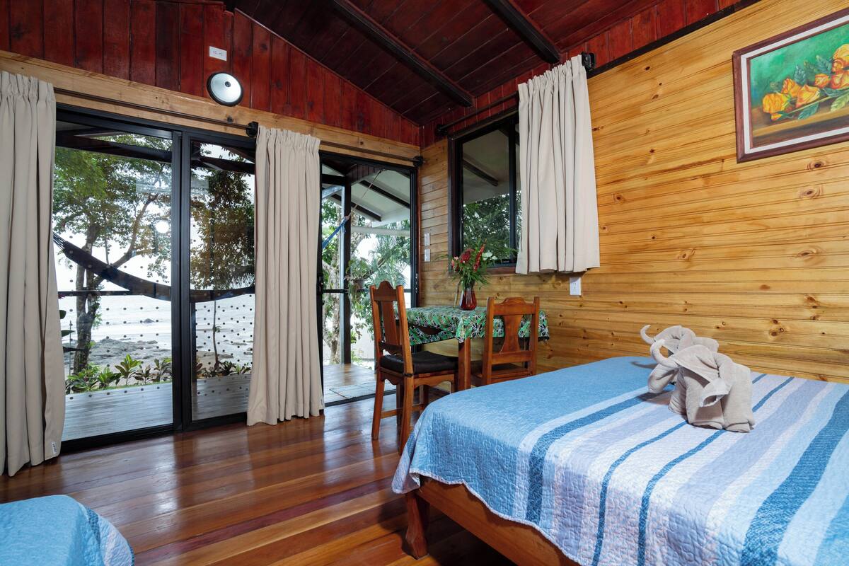 A room with wooden walls, a bed with a striped bedspread, a chair, a table with a green tablecloth, and a view of the beach through a glass door.