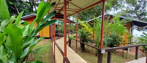 Una terraza de madera que conduce a un espacio cubierto con un letrero que dice 'BEACH', rodeado de vegetación exuberante y pequeñas construcciones.