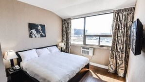 A hotel room with a large bed, a nightstand with a lamp, a painting on the wall, a window with curtains, and a flat-screen TV. - Nu Hotel Toronto (Mississauga)