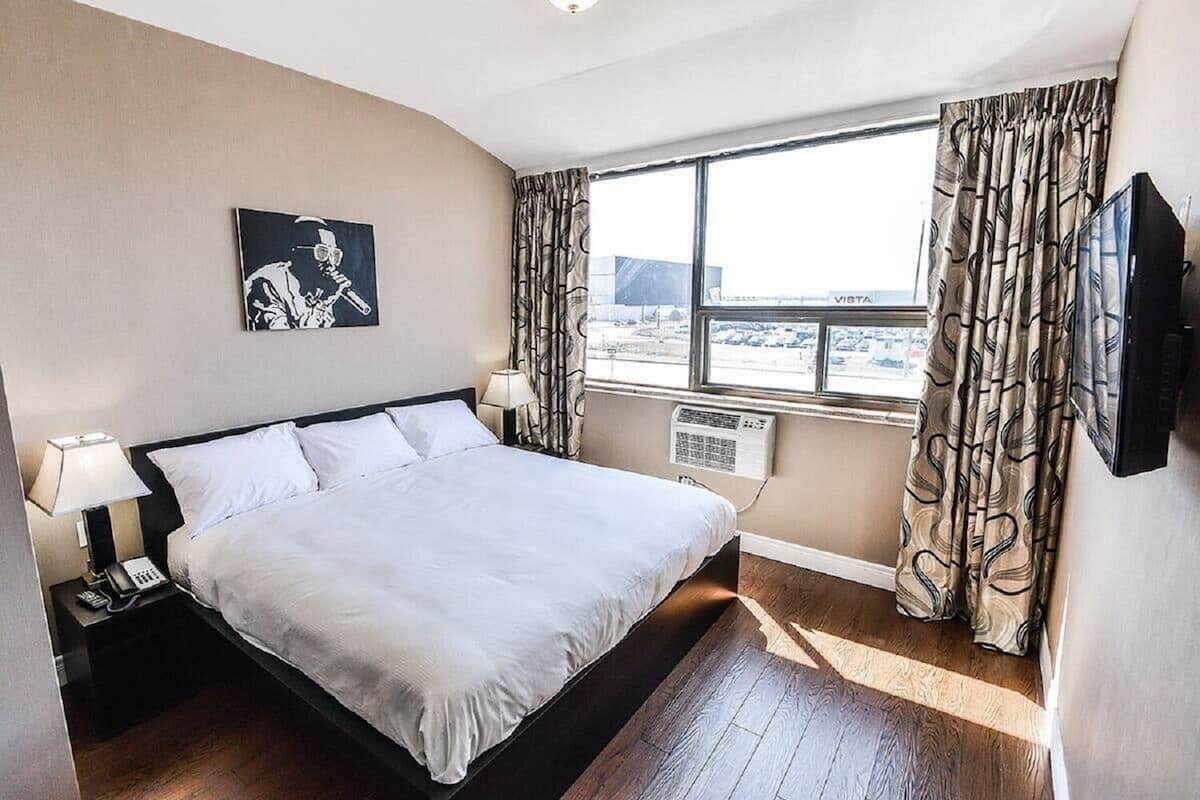 Hotellihuone, jossa on suuri sänky, yöpöytä lampun kanssa, taulu seinällä, ikkuna verhoilla ja litteänäyttöinen televisio.