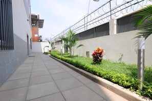 A narrow, paved pathway flanked by buildings with barbed wire on top, a metal pole, and a small garden with green shrubs and a red flower. - Inn Luanda (Luanda)