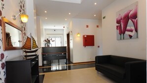 Eine Hotelhalle mit einer Rezeption, einer schwarzen Couch, einem Blumenbild und einer roten Notfallkiste.