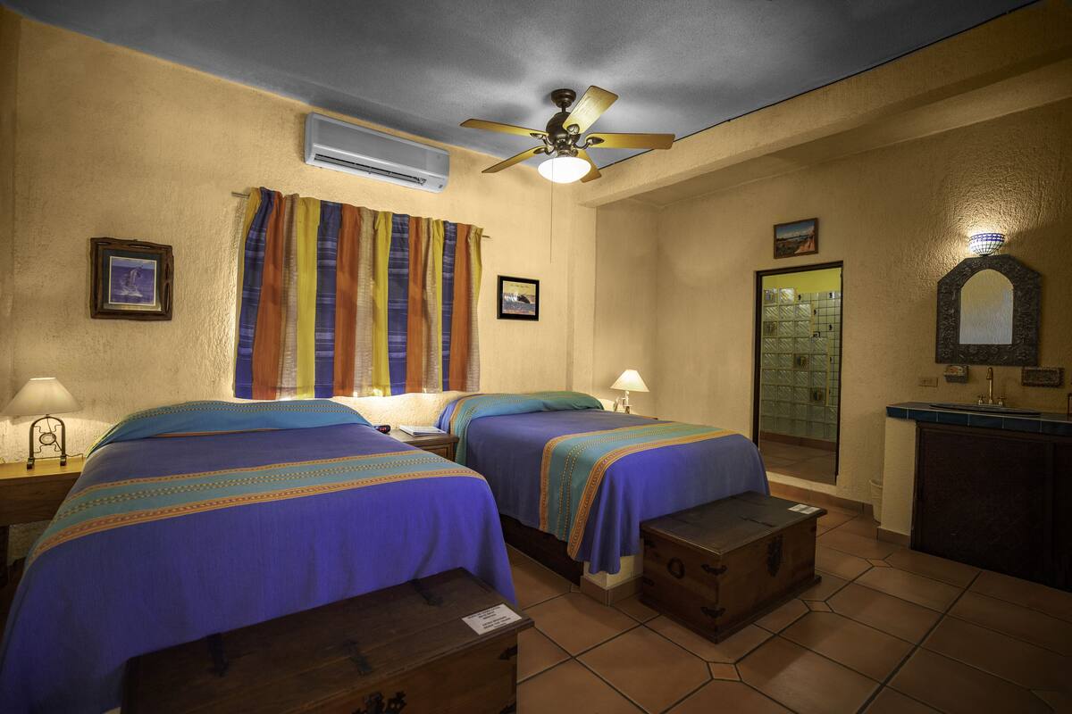 A hotel room with two beds, a ceiling fan, an air conditioning unit, a bathroom with a glass door, and a small desk with a lamp.