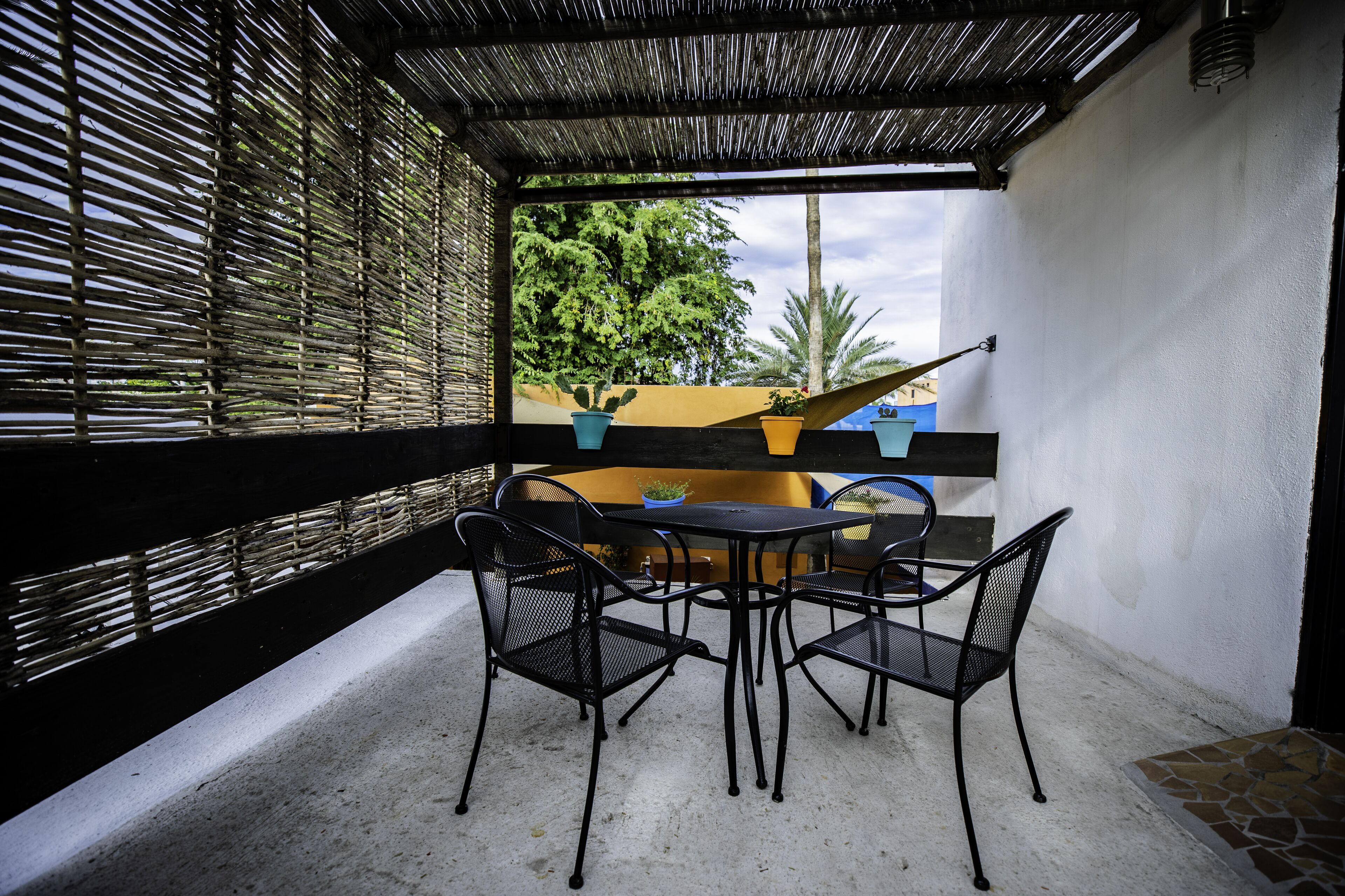 A covered patio with a table and chairs, a yellow wall, and potted plants.