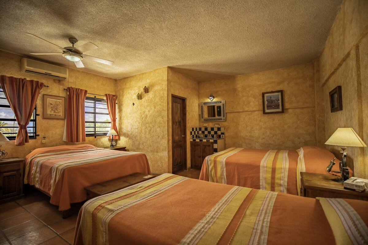 A hotel room with two beds, a ceiling fan, an air conditioning unit, a window with curtains, a nightstand with a lamp, and a checkered tablecloth on the wall.