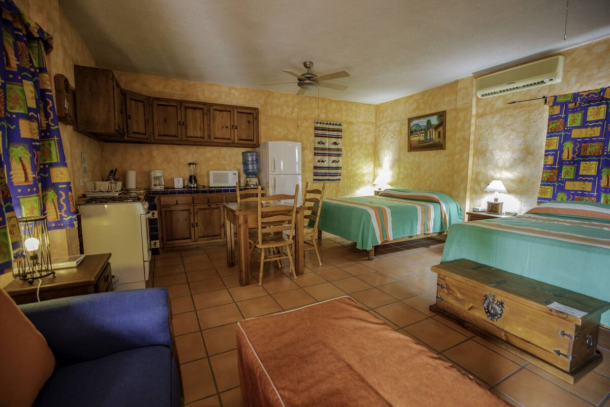 A compact room with a kitchenette, a bed with a green bedspread, a wooden chest, and a blue sofa.