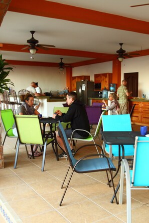 A dining area with chairs and tables, a kitchen in the background, and ceiling fans. - Posada Luna Sol (La Paz)