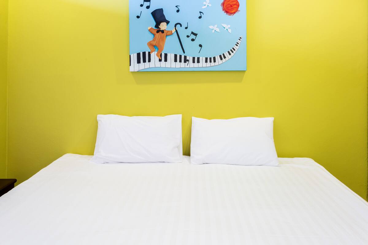 Ein Bett mit weißer Bettwäsche, zwei Kissen und eine Wand mit einem Gemälde, das eine Figur am Klavier zeigt.