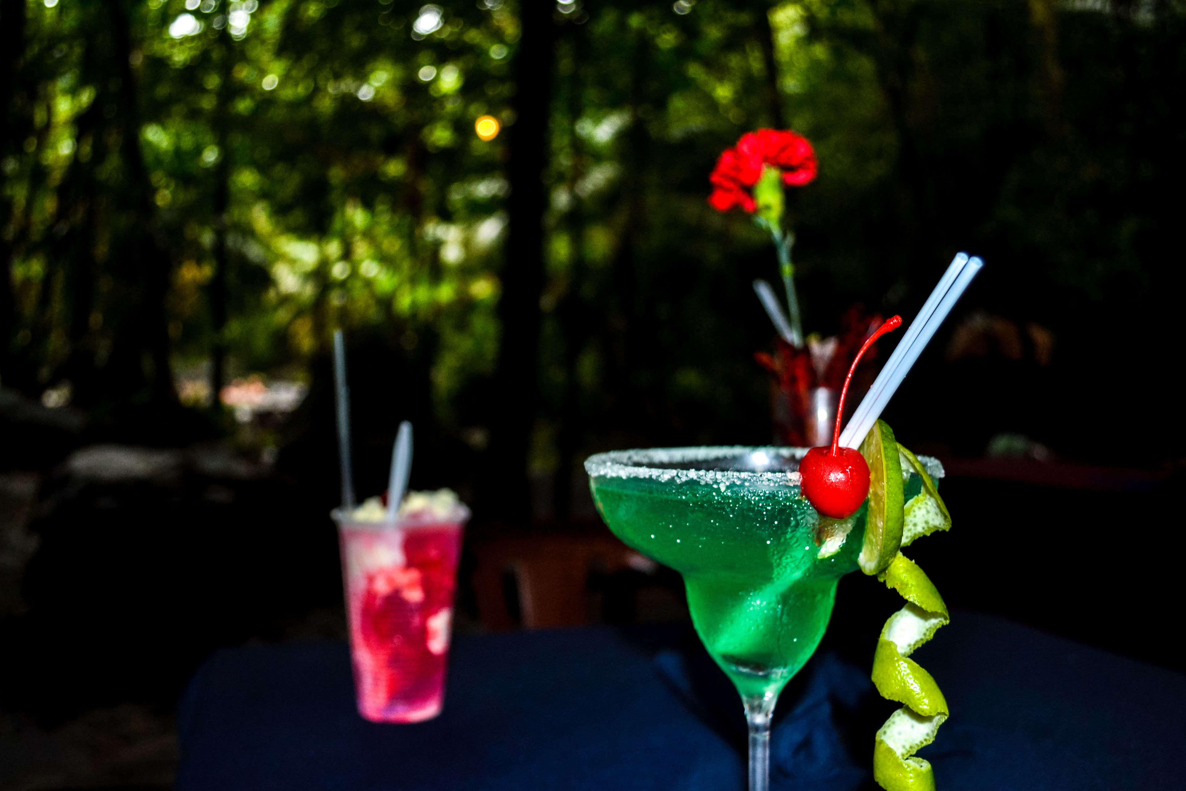 Zwei Cocktails auf einem Tisch mit dunklem Untergrund, einer mit einem grünen Getränk und Limettenscheibe, der andere mit einem roten Getränk und einer Kirsche.