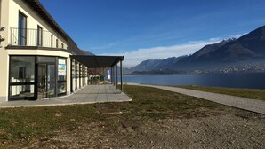 A modern building with large windows and a covered outdoor area, overlooking a lake and mountains. - Lake Como Beach Hostel (Domaso)