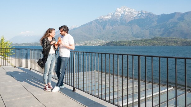 A couple standing on a waterfront promenade with a mountain backdrop. - Lake Como Beach Hostel (Domaso)