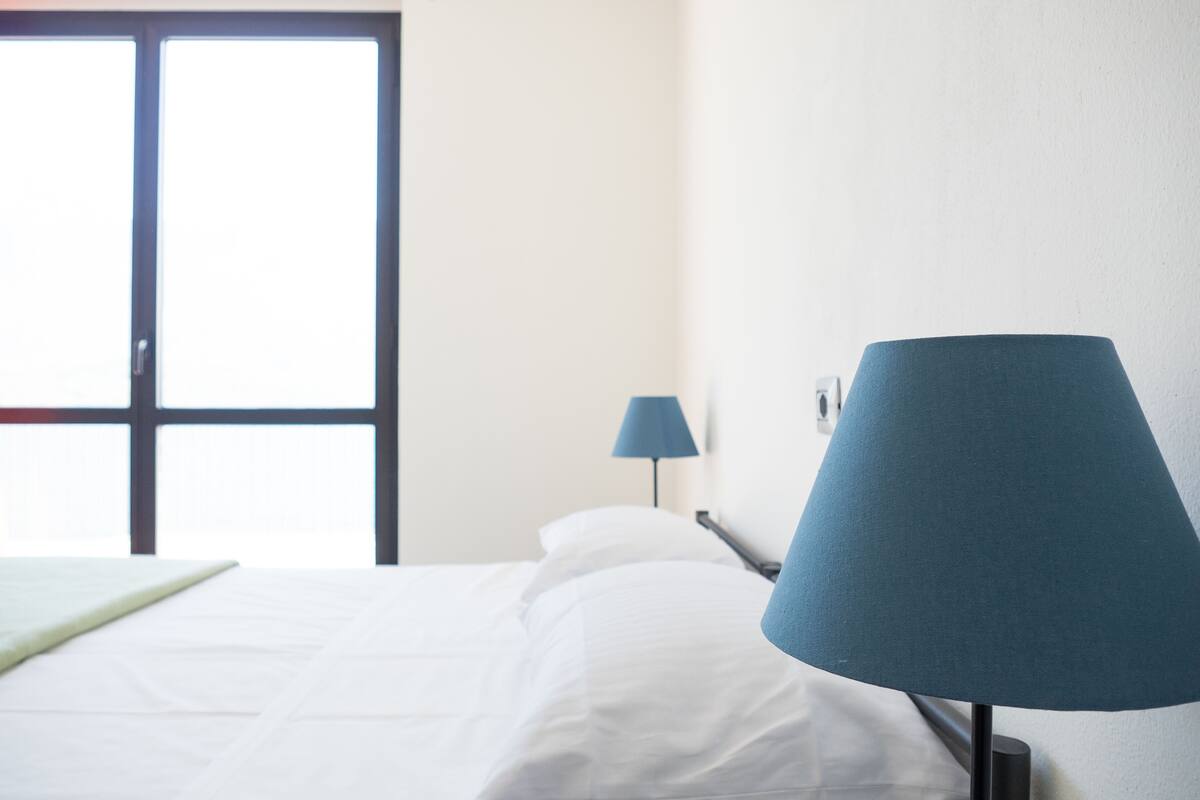 A hotel room with a bed, a blue lamp, and a window with blinds.